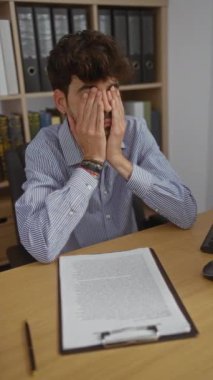 Ofiste hayal kırıklığına uğrayan İspanyol bir adam elleriyle, çalışma masasında kağıtlarla ve kalemle karışık, çizgili tişört giymiş.