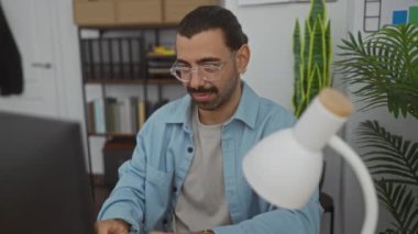 Ofiste masa başında çalışan genç İspanyol bir adam. Etrafta bitkiler ve ofis malzemeleri var.