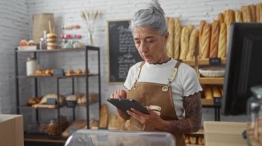 Fırında çalışan gri saçlı bir kadın, etrafında ekmek ve pastalar varken tablet kullanıyor. Kapalı alanda, işine odaklanmış ve profesyonel..