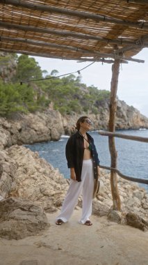 Caladeia, Mallorca 'da kadın, güneş gözlüğü ve plaj kıyafeti takıyor, bulutlu bir günde tahta bir gölgeliğin altında kayalık uçurumlar ve Akdeniz ile çevrili.