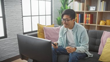 Modern bir oturma odasında televizyon izlerken uzaktan kumanda kullanan gülümseyen Asyalı bir adam..