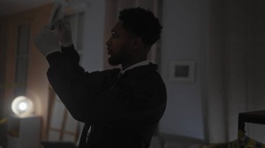 Odaklanmış bir Afro-Amerikalı, loş aydınlatılmış bir suç mahallinde, iş yerindeki bir dedektifi canlandıran kanıtları inceliyor..