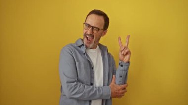 Orta yaşlı İspanyol bir adam gözlük takıyor zafer işareti yapıyor ve mutlu bir şekilde gülümsüyor izole edilmiş sarı arka planda.