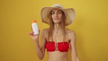 Bikinili kadın elinde güneş kremi tutuyor, parmaklarıyla işaret veriyor, gülümsüyor ve jest yapıyor mükemmel bir sembol, arkadaş canlısı ve neşeli, sarı arka planda izole edilmiş.