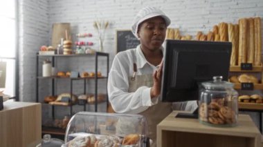 Önlük giyen bir kadın pastanede çalışıyor, hamur işi yapıyor ve parlak bir iç mekanda ekmek ve kurabiye sergiliyor.