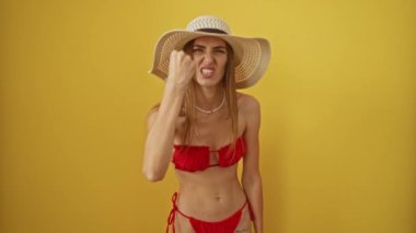 Bikini ve yaz şapkalı beyaz kadın izole edilmiş sarı arka planda öfkeli öfkeli yumruğuyla öfkeli görünüyor, agresif öfke ve hayal kırıklığını gösteriyor.