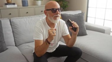 Gri saçlı adam, modern bir oturma odasında televizyon izlerken, elinde bir cep telefonu tutarak ve heyecanlı bir anın tadını çıkararak kutlama yapıyor..