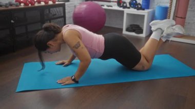 Genç bir kadın kapalı bir spor salonunda mavi bir paspasın üzerinde şınav çekiyor. İyi donanımlı bir spor salonunda atletizm yeteneklerini sergiliyor..