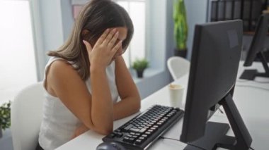 Ofisteki bilgisayarın başında çalışan bir kadın. Etrafı kapalı bitkiler ve dosyalarla çevrili. Hayal kırıklığı ve yorgunluk karışımı bir şey sergiliyor.
