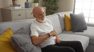 Gri saçlı adam evdeki rahat bir kanepede otururken mutlu bir şekilde dans ediyor. Yastık ve bitkilerle süslenmiş rahat bir oturma odasında eğleniyor..