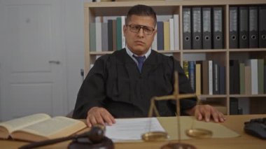 Masasında tokmak ve terazi olan yasal kitap ve belgelerle çevrili siyah cüppeli bir adam ofiste çalışıyor.
