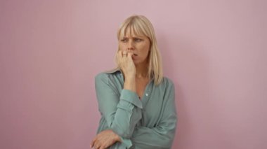 Endişeli bir kadın - orta yaşlı sarışın gergin bir şekilde duruyor, tırnaklarını yiyor ve stresli görünüyor, elleri dudaklarında, pembe arka planda.