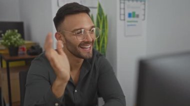 Genç bir adam, İspanyol asıllı, gülümseyen, gözlük takan, ofis çalışma alanında gözlük takan, kapalı, modern, teknoloji, bitkiler, arka planda masalar, çizelgeler, günlük kıyafetler.