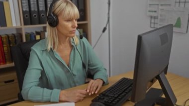 Orta yaşlı, sarı saçlı, kulaklık takan bir kadın. Ofis ortamında bilgisayarda çalışıyor. Arkasında bir sürü dosya ve kitap var.