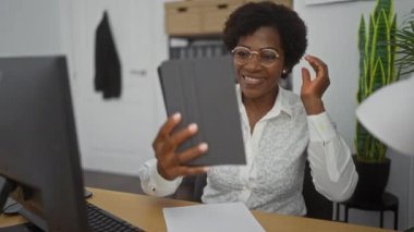 Orta yaşlı, kıvırcık saçlı bir kadın ofis ortamında tabletle selfie çekiyor. Ofis mobilyaları ve belgelerle dolu kapalı bir ortamda yaşıyor.