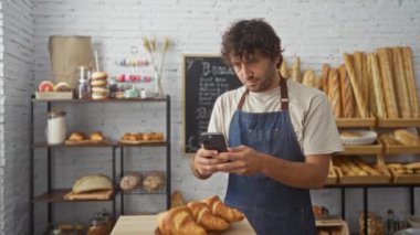 Fırında genç bir adam, yeni pişmiş kruvasanların fotoğrafını çekiyor. Telefonu evde, ekmek ve hamur işleriyle çevrili.