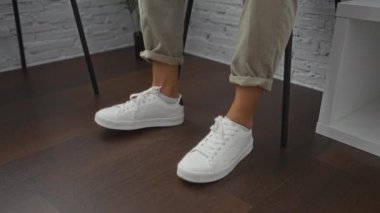 Beyaz spor ayakkabı ve haki pantolon giyen bir adam modern bir odada ayakları çaprazlanmış, karanlık bir ahşap odada bekler..