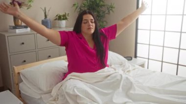 Pembe tişörtlü genç bir kadın, büyük pencerelerden süzülen bitkiler ve ışıkla, rahat bir yatak odasında sabah meditasyonu yapıyor.