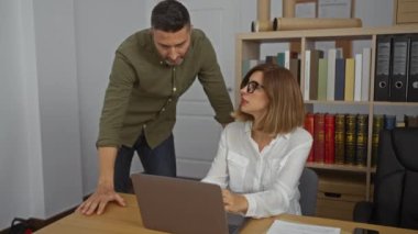 Kadın ve erkek, arka planda dizüstü bilgisayarı ve kitaplığı olan bir ofiste birlikte çalışıyor. İş ortamında iş arkadaşı gibi görünüyorlar..