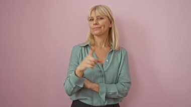 Orta yaşlı bir kadın, sarışın, tişört giyiyor, ayakta duruyor ve mutlu bir parmakla işaret ediyor ve pembe bir arka plan üzerinde gülümsüyor.