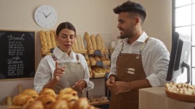 Fırında konuşan erkek ve kadın fırıncılar etrafları taze ekmek ve pastalarla çevrili, raflar ve arka planda menü tahtasıyla.