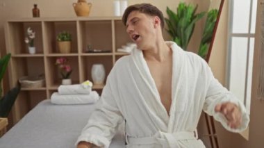 Beyaz cüppeli genç adam huzur dolu bir spa odasında dinleniyor. İçinde bitki ve raflar var. Huzurlu bir sağlık ortamı yaratıyor.