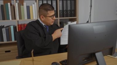 Kitaplık ve masaüstü monitörlü bir ofiste video görüşmesinde elinde bir belge tutan İspanyol asıllı genç bir adam..