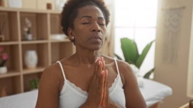 Kaplıca odasında meditasyon yapan bir kadın, elleri kapalı, gözleri kapalı, kolsuz tişört giyen, rafları olan ve arka planda kapalı bitkiler olan bir kadın.