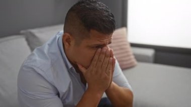 Elleri kapalı evde oturan genç İspanyol bir adam stres jesti ile yüzünü kapatıyor ya da evdeki bir kanepede endişeleniyor.