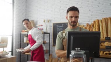 İki adam bir pastanede birlikte çalışıyor. Ekmek ürünleri sergileniyor. Biri kasayı işletiyor, diğeri de fırınlanmış mallara bakıyor.