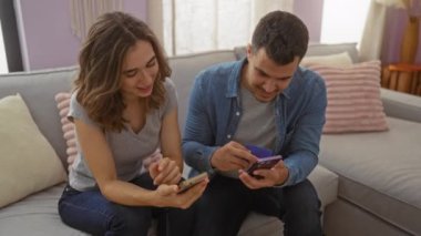 Çift, rahat bir oturma odasında kanepede otururken akıllı telefonlar ve kredi kartı kullanıyor, gülümsüyor ve evde birlikte vakit geçiriyorlar.