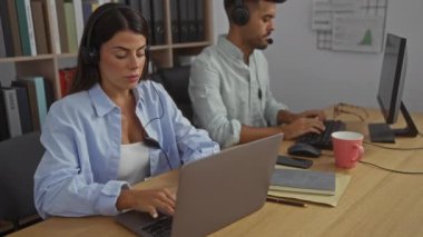 Ofis masasında çalışan kadın ve erkek, kulaklıklarla birlikte, ofis ekipmanlarıyla çevrili, kapalı bir ortamda, profesyonel bir çalışma ortamını işaret ediyor..