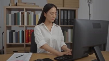 Ofiste çalışan, arka planda kitap rafları olan, gözlüklü, beyaz gömlekli, odaklanmış ve profesyonel görünen bir kadın.