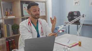 Klinikteki genç bir İspanyol erkek veteriner akıllı telefonunu elinde dizüstü bilgisayar, stetoskop ve ilaçla masada otururken kullanıyor..