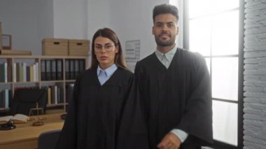 Kadın ve erkek, kitaplıkların ve pencerelerin önünde kolları çaprazlanmış yargıç cüppesi giyerek ofiste yan yana duruyorlar.