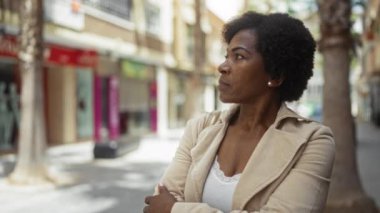 Bir şehir ortamında kolları çapraz duran kadın Afro-Amerikan kıvırcık saçlarını ve zarif bej ceketini sergiliyor.
