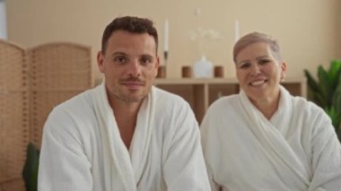 Anne ve oğlu bir kaplıcada oturuyor, beyaz elbiseler giyiyor, gülümsüyor, sağlık hizmetlerinden keyif alıyor, modern, kapalı bir salon ortamında rahatlıyor.