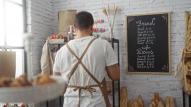 Genç adam pastanede taze kruvasan paketliyor. Kahverengi bir önlük giyerken mutlu bir şekilde gülümsüyor. İçi çeşitli fırınlanmış ürünlerle dolu raflarla ve karton menülerle dolu..