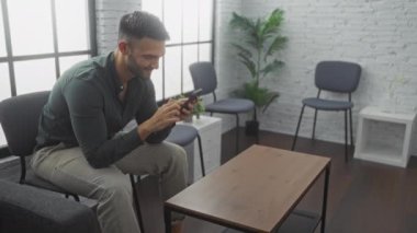 Sakallı genç bir İspanyol adam kapalı bir bekleme odasında ya da lobide oturuyor, cep telefonunu kullanıyor, gülümsüyor ve mutlu görünüyor..