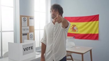 İspanyol asıllı genç adam ayakta dikilip parmağıyla işaret ediyor. İspanyol seçim okulunda kameraya özgüvenli ve ciddi bir jest yapıyor.