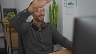 Gözlüklü ve sakallı genç bir İspanyol modern ofis ortamında kulaklık takarken mutlu mesut çalışıyor..