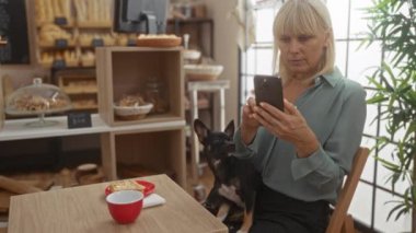 Orta yaşlı bir kadın fırında chihuahua ile oturuyor, telefonlarını karıştırıyor, hamur işleri ve kahve yapıyor.