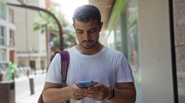 Şehir caddesinde akıllı telefon kullanan genç adam, eli başının üstünde ve çantası omzunda düşünceli görünüyor.