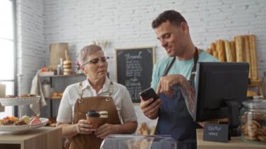 Kadın ve erkek, ekmek ve hamur işleriyle bir pastanede birlikte çalışıyorlar, akıllı telefon kullanıyorlar ve tezgahın arkasında duruyorlar.