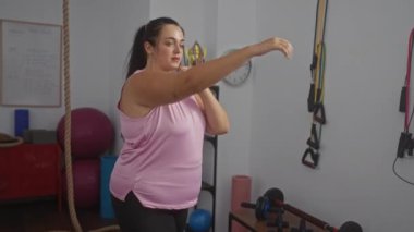 Arka planda halatlar ve spor aletleriyle egzersiz yapan, pembe bir atlet giyen ve boks hareketleri sergileyen bir kadın..