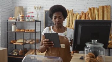 Fırında çalışan bir kadın elinde tabletle çeşitli ekmek, hamur işleri ve kavanozlardaki kurabiyelerle dolu bir tezgahın üzerindeki bilgisayar ekranına bakıyor.
