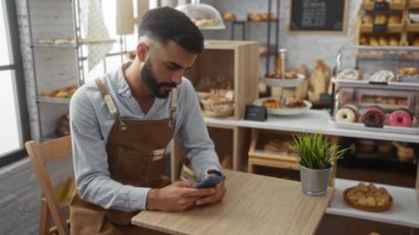 Masada oturan sakallı ve önlüklü genç adam ekmek ve hamur işleri sergilenen bir fırında akıllı telefon kullandı.