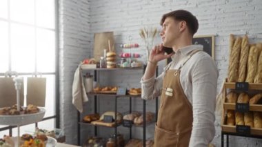 Fırında konuşan genç adam ekmek rafının ve pencerenin yanında önlük takıyor.