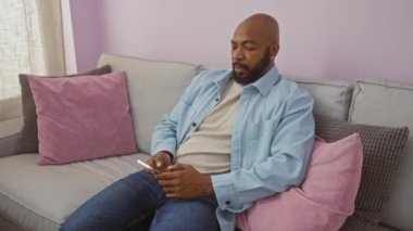 Oturma odasındaki kanepede pembe ve gri yastıklı akıllı telefon kullanırken esneyen genç adam.