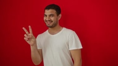 Beyaz tişört giyen, mutlu bir yüzle gülümseyen, göz kırpan, iki parmağı izole edilmiş kırmızı arka planda zafer işareti yapan İspanyol genç adam.
